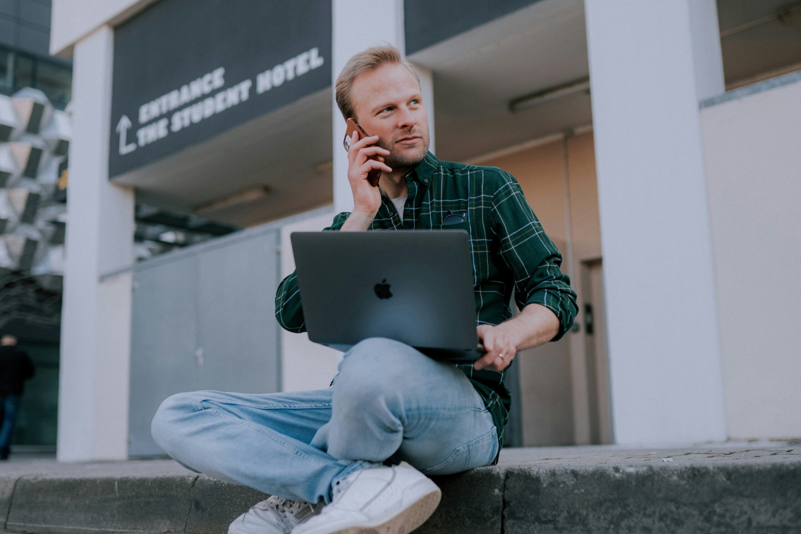 man in green and black plaid dress shirt and blue denim jeans sitting on gray concrete